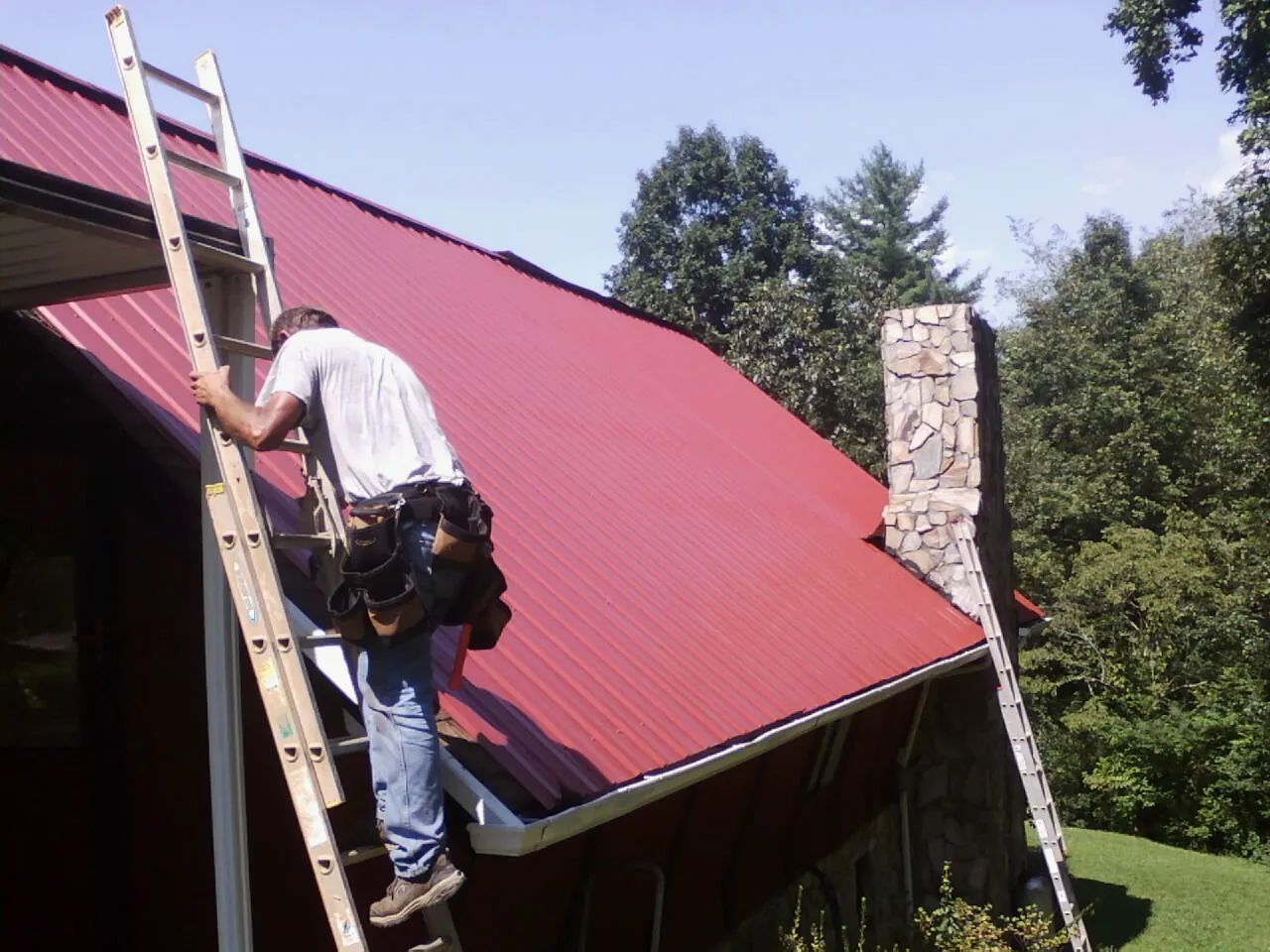 Roofer on a ladder beside a red metal roof and stone chimney — Soffit & Fascia Repair by Roofing Brothers in Metuchen